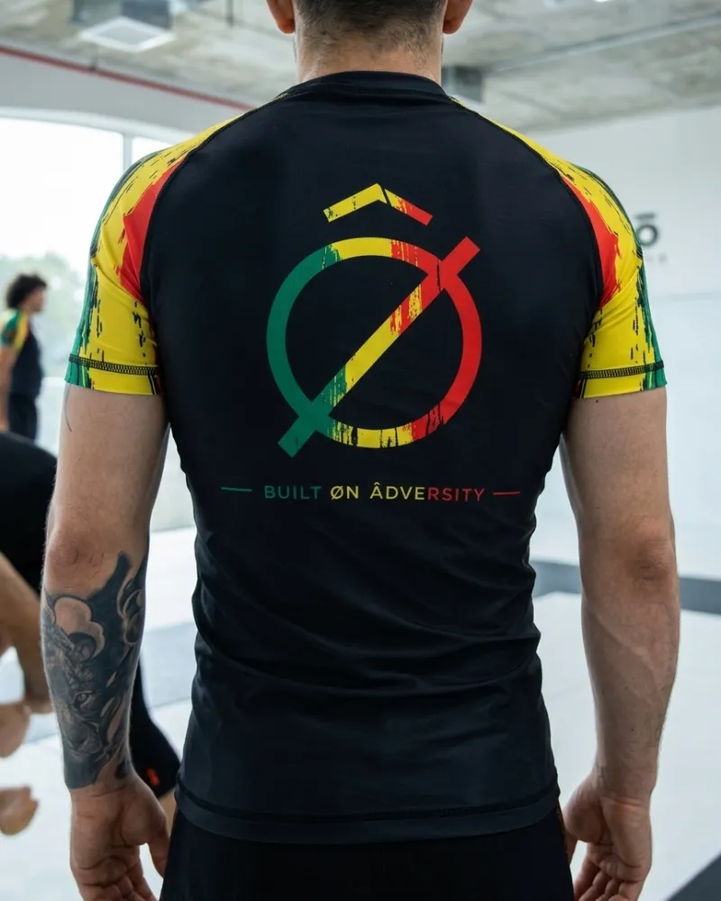Espalda de un rashguard negro con mangas verdes, amarillas y rojas, el símbolo de BÔA Collective y la frase Built on Adversity
