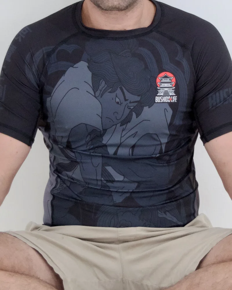 Rashguard negro de manga corta con la ilustración de un luchador de jiu jitsu y el logo Bushido Life, puesto por un hombre sentado en posición de loto